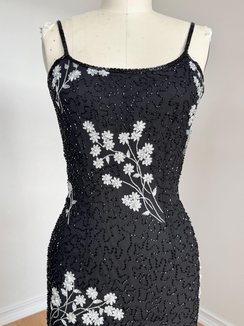 May include: A black beaded dress with white floral embroidery. The dress features thin spaghetti straps and a fitted silhouette. The floral design is concentrated on the bodice and lower portion of the dress.