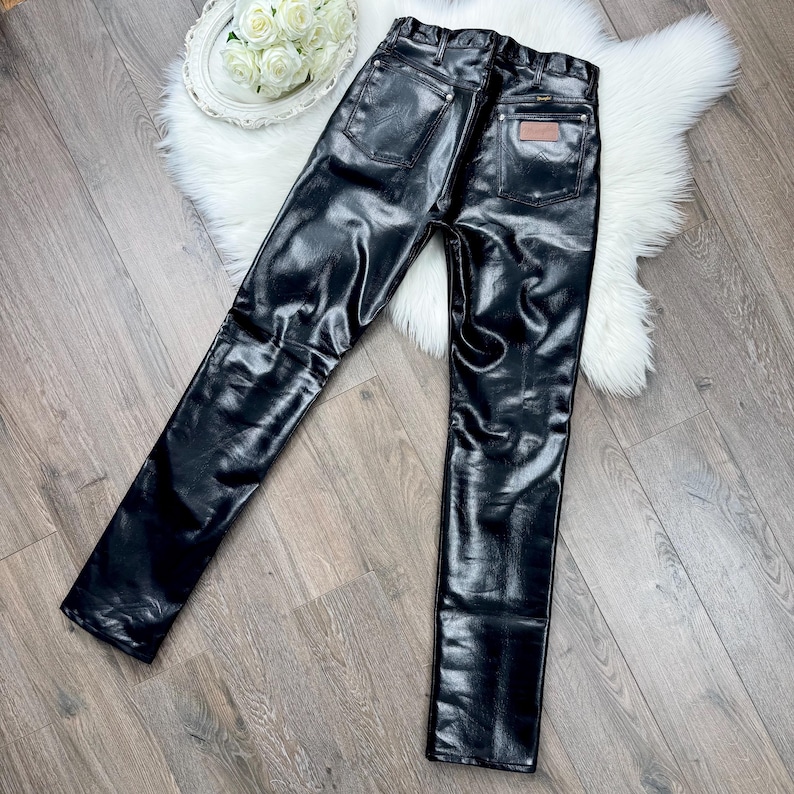 May include: Black, shiny faux leather trousers with a classic five-pocket design. The trousers have a high waist and a straight leg. A small, rectangular patch is visible on the back pocket. The trousers are displayed on a wooden surface.