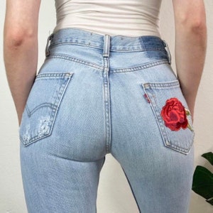 May include: Light blue denim jeans with a red embroidered rose on the back pocket.