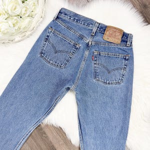 May include: Blue denim jeans featuring a high-waisted cut, two back pockets with arcuate stitching, a tan leather patch on the waistband, and a small red tab on the right pocket. The jeans are constructed with durable cotton denim fabric.