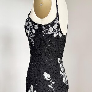 May include: A black beaded evening dress with white floral embroidery. The dress features a scoop neckline, spaghetti straps, and a fitted silhouette. The floral design is concentrated on the bodice and along the side seams.