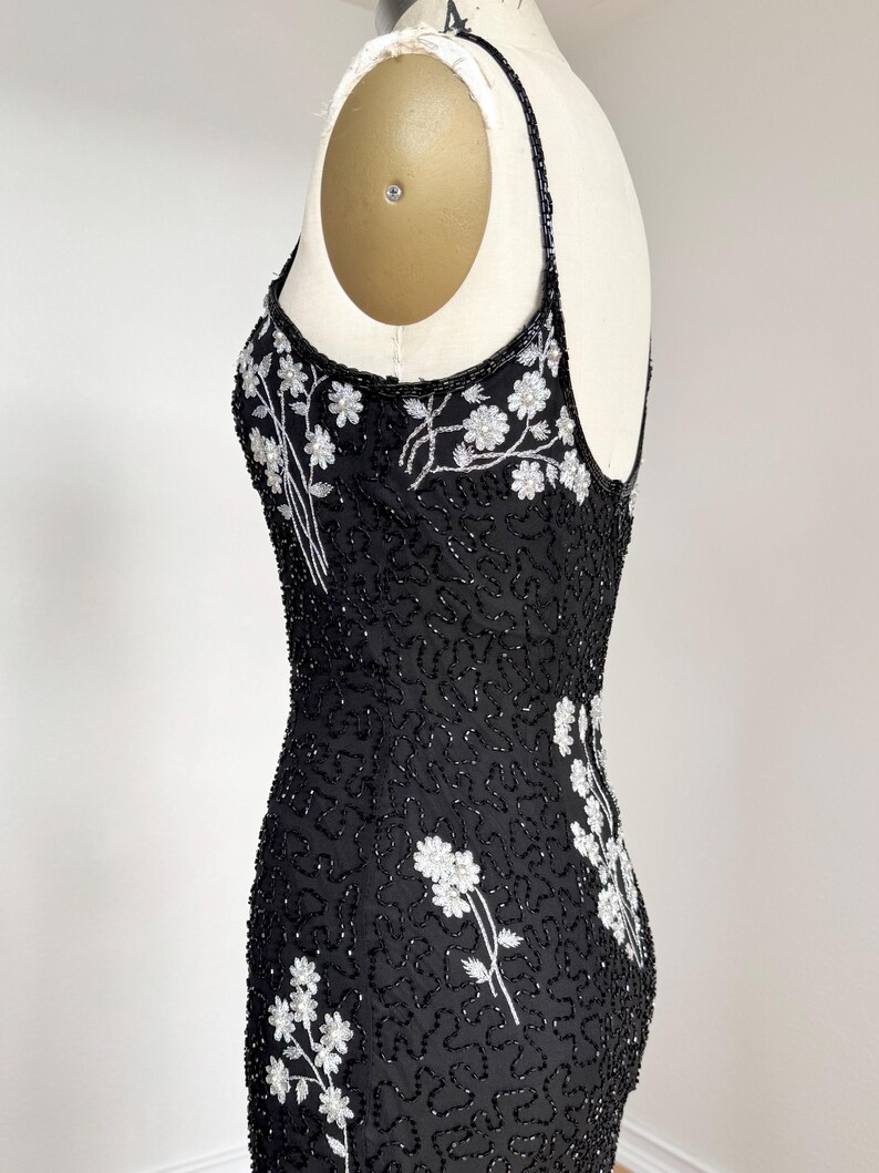 May include: A black beaded evening dress with white floral embroidery. The dress features a scoop neckline, spaghetti straps, and a fitted silhouette. The floral design is concentrated on the bodice and along the side seams.