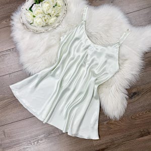 May include: A light green satin slip dress with spaghetti straps. The nightgown has a subtle sheen and a delicate embroidered detail near the neckline. The dress is draped over a fluffy white faux fur rug, with a decorative tray of white roses in the background.
