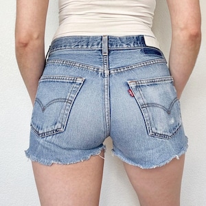 May include: Light blue denim shorts with a frayed hem. The shorts have classic five-pocket styling, a button and zipper closure, and the iconic Levi's red tab on the back pocket. The shorts are worn by a person.