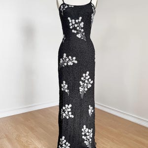 May include: A black, floor-length evening gown with spaghetti straps. The dress is embellished with white floral embroidery. The dress is displayed on a mannequin against a neutral background. The dress is made of a beaded fabric.