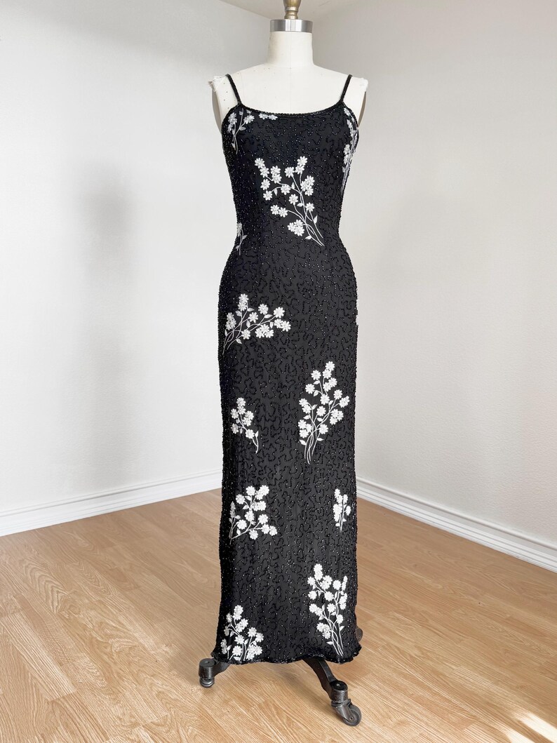 May include: A black, floor-length evening gown with spaghetti straps. The dress is embellished with white floral embroidery. The dress is displayed on a mannequin against a neutral background. The dress is made of a beaded fabric.