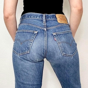 May include: Blue denim jeans featuring a high-waisted fit, two back pockets with arcuate stitching, a tan leather-like patch on the waistband, and a small red tab on the right pocket. The jeans are worn with a plain black top.
