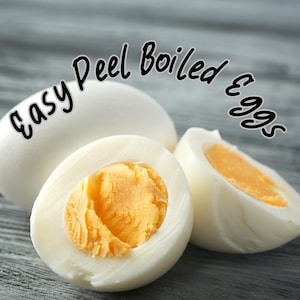 Op de afbeelding: Afbeelding van drie gekookte eieren, waarvan één heel en twee helften, met heldergele dooiers. De tekst "Easy Peel Boiled Eggs" is in een cursief lettertype over de bovenkant van de afbeelding geschreven. De eieren liggen op een grijze houten ondergrond.