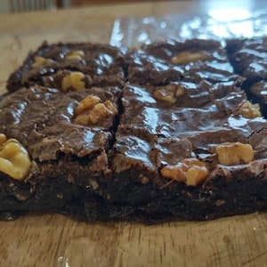 May include: Close-up of several square brownies with a shiny, dark chocolate surface. Each brownie is topped with pieces of walnuts. The brownies are arranged on a wooden surface, suggesting a homemade dessert.