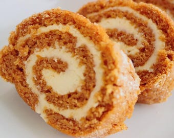 May include: Close-up of sliced pumpkin roll with a creamy white filling. The cake has a light brown color and is dusted with a fine powder. The filling swirls within the roll, creating an appealing visual texture. The dessert is on a white surface.