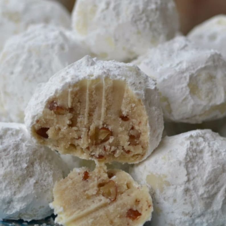 Puede incluir: Primer plano de un plato de bolas de nieve, un tipo de galleta, espolvoreada con az&uacute;car glas. Algunas de las bolas de nieve est&aacute;n cortadas por la mitad, revelando un centro relleno de nueces de pec&aacute;n.