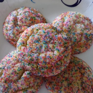 May include: Four round sugar cookies covered in multicolored sprinkles arranged on a white paper plate with a blue and purple swirl pattern. The cookies have a cracked, textured surface and are coated in red, blue, green, and yellow sugar.