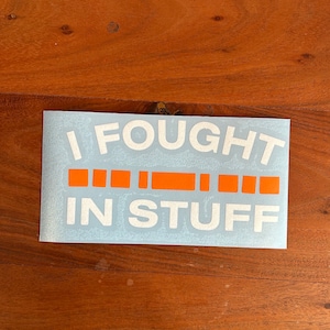 May include: A white vinyl sticker with the text "I FOUGHT IN STUFF" in white letters. The word "FOUGHT" is in a larger font size than the rest of the text. There are six orange stripes below the word "FOUGHT".