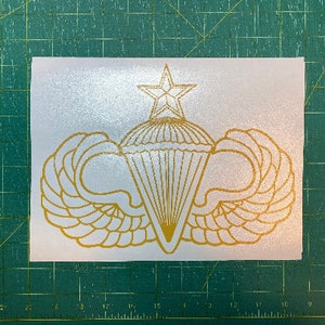 May include: Gold-colored vinyl decal of a paratrooper badge with a star and wings. The badge is on a white background.