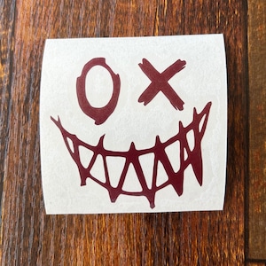 May include: A burgundy red vinyl sticker with a stylized smiling face with a single eye and an 'X' over the other eye. The mouth is a jagged, sharp smile.