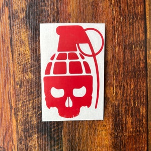 May include: Red vinyl sticker of a skull with a grenade on top. The skull has a menacing expression with two eye sockets and a large, open mouth.