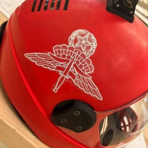 May include: A bright red helmet with a white emblem featuring wings, a star, and crossed swords. The helmet has a black visor and a black strap. Black stripes are visible on the top of the helmet.