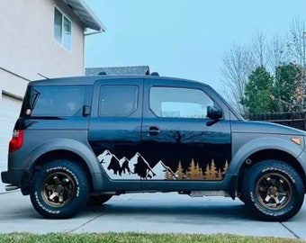 May include: A black Honda Element SUV with a mountain and forest decal on the side. The decal is in white, black, and gold. The car is parked on a paved driveway.