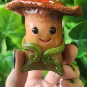 Op de afbeelding: Een klein, antropomorf paddenstoelfiguurtje met een glanzende rode hoed met witte stippen. Het beige, cilindervormige lichaam heeft een lachend gezicht met zwarte ogen en twee groene, rankachtige armen die aan de voorzijde zijn gekruld en eindigen in puntige voeten.