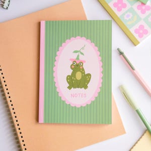 Peut inclure: Un carnet vert avec un dos rose et une illustration de grenouille. La grenouille porte un chapeau rose et se trouve à l'intérieur d'un ovale rose avec le mot "NOTES" en bas. Le carnet est posé sur un carnet de couleur pêche.