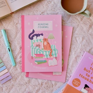 May include: A pink reading journal with the words "READING JOURNAL" and a cat illustration. A pen and sticky notes are to the left. A cup of tea and books are also in the image.