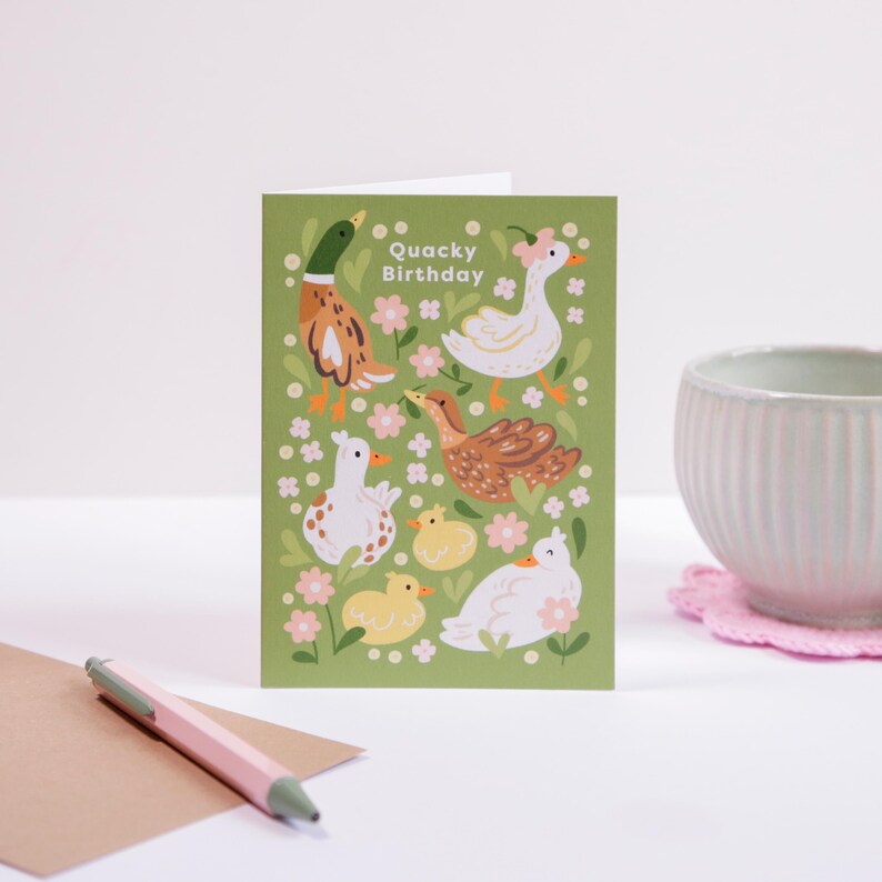 Cute illustrated duck birthday card with playful ducks and pink flowers on a green background, featuring the text 'Quacky Birthday', styled beside a pen, envelope, and pastel cup.
