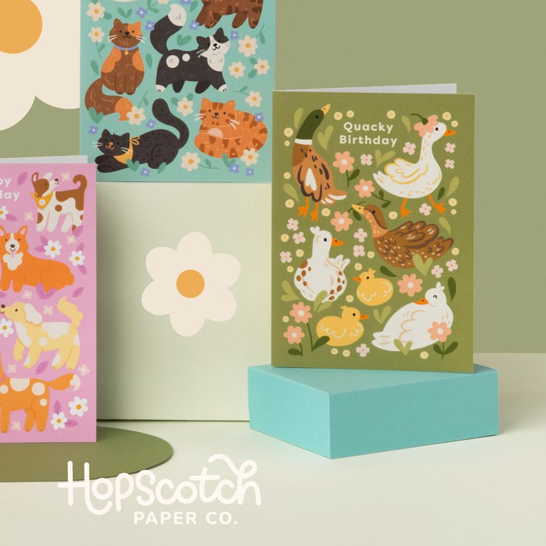Display of illustrated animal birthday cards by Hopscotch Paper Co, including a duck-themed ‘Quacky Birthday’ card, styled with cat and dog designs in a pastel setting.