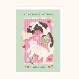 May include: Greeting card with the text "I LOVE DOING NOTHING WITH YOU." The illustration features two cats, one black and white, the other white, surrounded by hearts, a book, and a video game controller on a pink striped background.