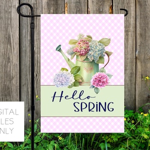 May include: A garden flag with a pink gingham background and a green watering can filled with pink and blue hydrangeas. The words "Hello SPRING" are written in navy blue. The flag is displayed on a black metal stand.