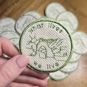 Puede incluir: Un parche redondo blanco roto con texto y diseño bordados en verde. El texto dice "what lives we live". El parche tiene un borde verde y una apariencia de tela texturizada. Varios parches similares son visibles en el fondo.