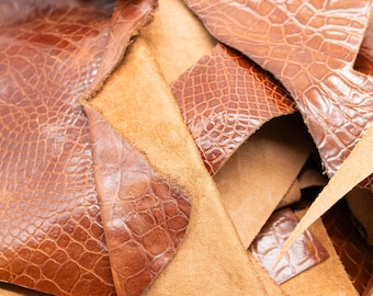 May include: A collection of brown leather scraps featuring a textured crocodile skin pattern on the top surface and a soft, tan suede texture on the underside. The pieces are irregularly shaped with visible raw edges and varying sizes.