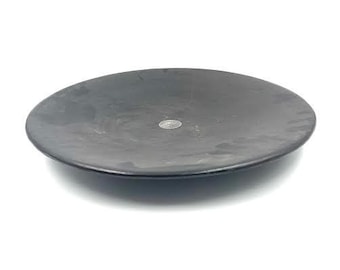 May include: A round, dark gray plate with a slightly textured surface. A small, silver coin is visible on the plate's surface. The plate is set against a white background.