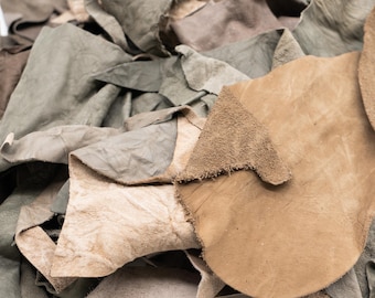 May include: A collection of irregular leather scraps in various shades of brown, tan, and muted olive green. The pieces feature diverse textures, ranging from smooth surfaces to rough, unfinished edges, layered together in a dense, overlapping pile.