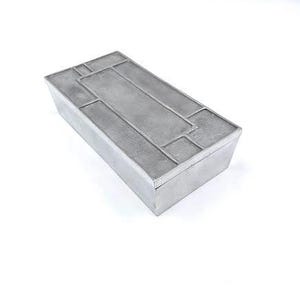 Puede incluir: Una caja rectangular de plata con un diseño geométrico en la tapa. La caja está hecha de metal y tiene un acabado liso y pulido.