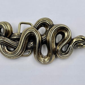 May include: A gold-toned metal belt buckle shaped like a snake. The snake is curled and intertwined, with a detailed, textured surface.