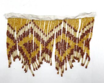 May include: A fringe trim with a geometric pattern in shades of brown and yellow. The fringe is made of small beads and is attached to a white fabric backing.