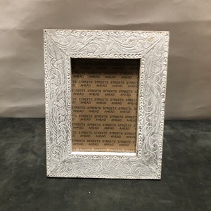 Puede incluir: Marco de fotos de madera blanco con un diseño tallado ornamentado. El marco tiene una abertura rectangular y está decorado con un patrón repetitivo de hojas y espirales. El marco está vacío.