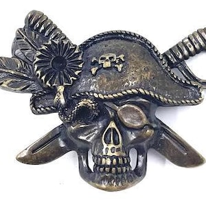 May include: A bronze pirate skull belt buckle with a feathered hat, an eyepatch, and crossed swords. The skull has a detailed design with a bone structure and a crossbones on the hat.