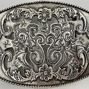 May include: A silver-toned metal belt buckle with a decorative, intricate design featuring a floral pattern and swirls. The buckle has a rope-like border.