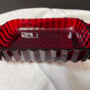 May include: A rectangular, ruby-red glass dish with a geometric design. The dish has a zig-zag edge and handles on each end. The interior features a diamond pattern. The dish is likely vintage and suitable for serving or display.