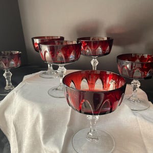May include: Six ruby red champagne coupe glasses with clear stems and bases. The glasses have a decorative cut pattern around the bowl. The glasses are arranged on a white cloth.