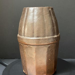 May include: A brown, barrel-shaped ceramic container with a ridged top and a geometric, faceted lower section. The container has a dark, earthy tone and a slightly rough texture, suggesting a rustic or vintage aesthetic. The container is likely used for storage or display.