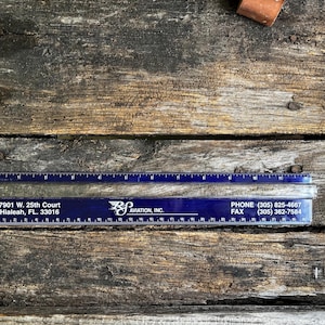 May include: A clear, cylindrical glass rod positioned against a blue plastic ruler. The ruler displays the text 7901 W. 25th Court, Hialeah, FL. 33016, RS AVIATION, INC., and phone numbers (305) 825-4667 and (305) 362-7584. The rod measures 12 inches long.