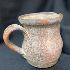May include: A handmade ceramic mug with a rounded body and a sturdy handle. The mug features a speckled, earthy brown glaze with hints of blue and orange around the rim and handle. The mug is designed for hot beverages.