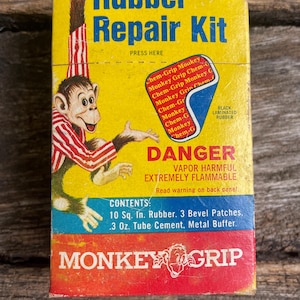 May include: A rectangular yellow, blue, and red box for a Rubber Repair Kit. The box features a cartoon monkey in a red and white striped shirt, the text "Rubber Repair Kit", "DANGER", "VAPOR HARMFUL", "EXTREMELY FLAMMABLE", and "MONKEY GRIP".