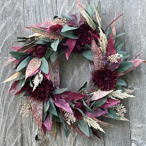 May include: A circular wreath featuring deep burgundy dahlia flowers, dark green eucalyptus leaves, and variegated foliage with reddish-brown speckles. The arrangement includes small clusters of cream-coloured dried grasses and textured botanical accents.