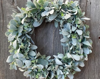 May include: A green and gray artificial wreath made of lambs ear leaves. The wreath is hanging on a wooden door.