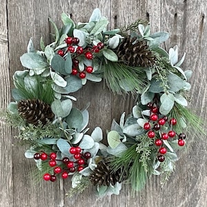 May include: A circular wreath featuring faux greenery, red berries, and pinecones. The wreath is composed of gray-green leaves, with accents of red berries and brown pinecones. The wreath is displayed against a weathered wooden background.