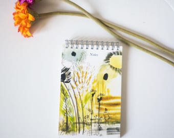 May include: A white spiral bound notebook with a colourful floral design and the word "Notes" printed on the cover. The notebook is sitting on a white surface with two pink and orange flowers.