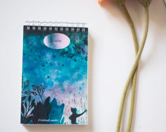 May include: A blue and green watercolour notebook with the word "Notes" on the cover. The notebook has a spiral binding and a silhouette of a person reaching up towards the sky. The notebook is on a white surface with two flowers.
